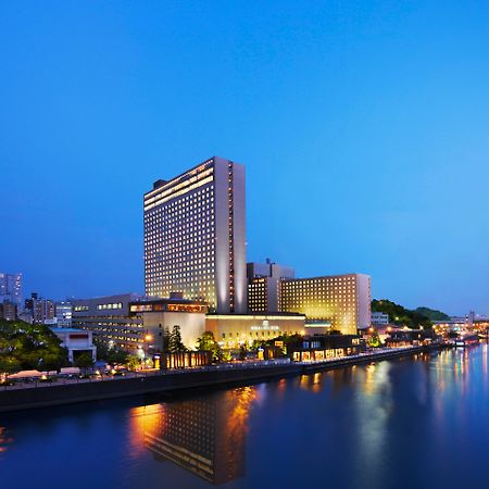 Rihga Royal Hotel Osaka, Vignette Collection By Ihg Zewnętrze zdjęcie The photo shows a large hotel building situated near a body of water, likely a river. The scene is illuminated by soft evening lighting, giving the environment a tranquil, serene feel. In the background, there are various buildings and structures, su