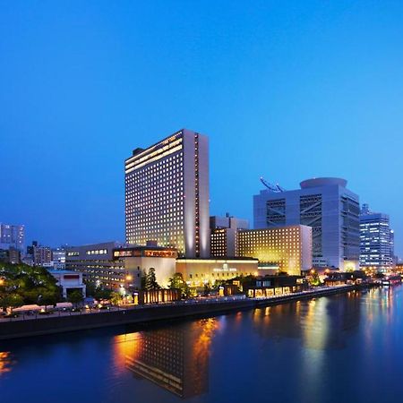 Rihga Royal Hotel Osaka, Vignette Collection By Ihg Zewnętrze zdjęcie The photo shows a cityscape at dusk, featuring a modern hotel and several tall buildings along a river. The structures are illuminated with lights, reflecting beautifully on the water. The atmosphere has a serene and urban feel, with a clear blue sky