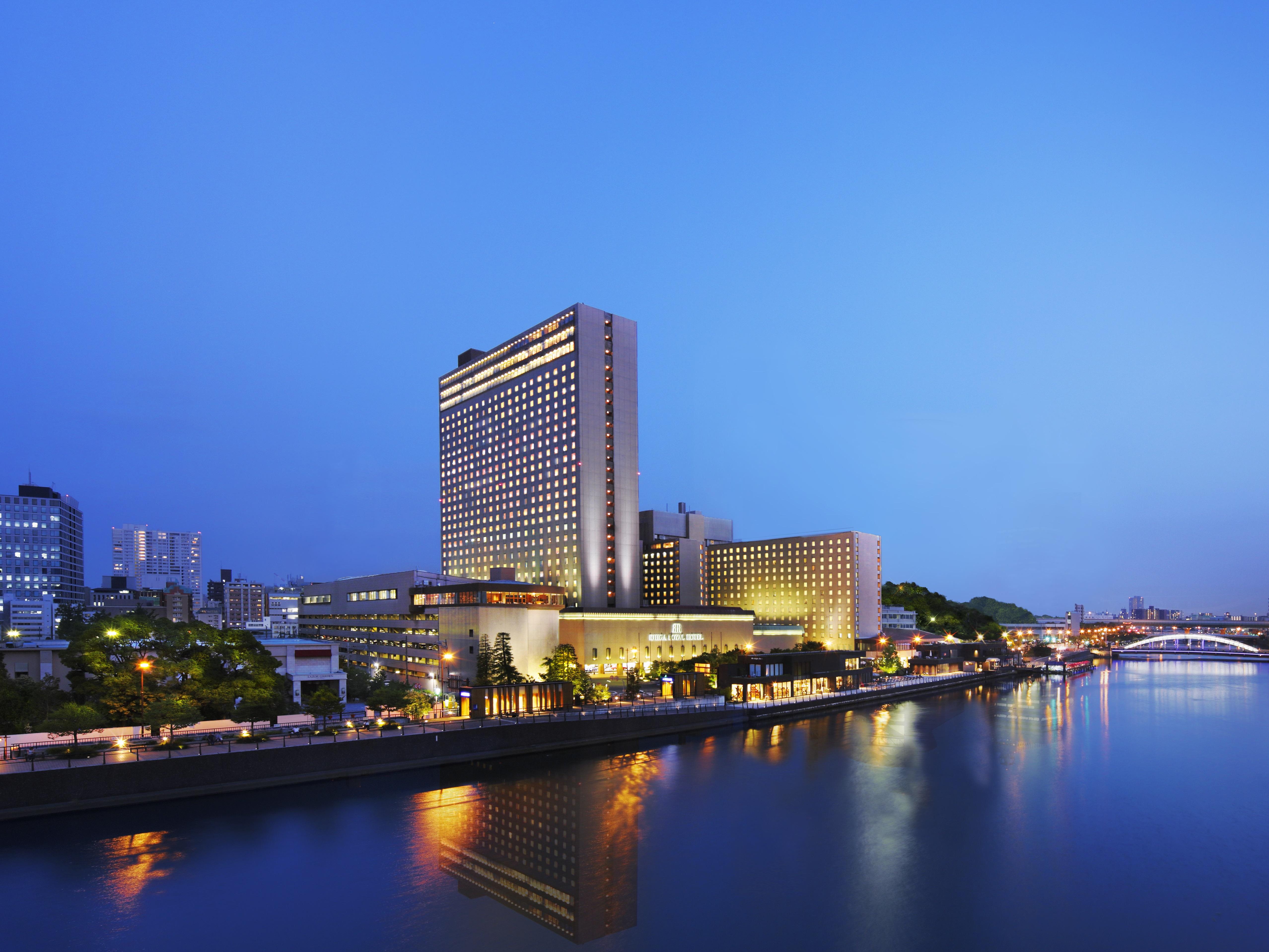 Rihga Royal Hotel Osaka, Vignette Collection By Ihg Zewnętrze zdjęcie The photo shows a large hotel building situated near a body of water, likely a river. The scene is illuminated by soft evening lighting, giving the environment a tranquil, serene feel. In the background, there are various buildings and structures, su