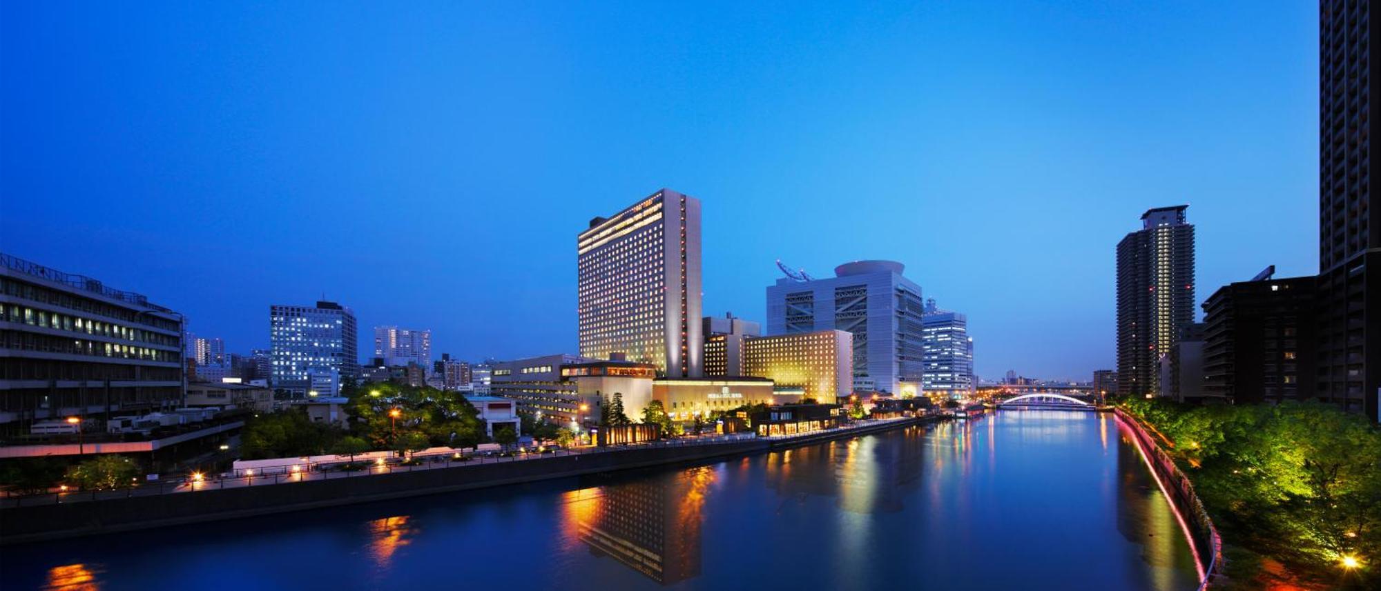 Rihga Royal Hotel Osaka, Vignette Collection By Ihg Zewnętrze zdjęcie The photo shows a cityscape at dusk, featuring a modern hotel and several tall buildings along a river. The structures are illuminated with lights, reflecting beautifully on the water. The atmosphere has a serene and urban feel, with a clear blue sky